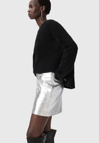 Melns pūkains džemperis kombinācijā ar spīdīgu sudraba mini svārku un melnām zābakiem līdz ceļiem. Modele pozē sāniski gaišā fona priekšā.