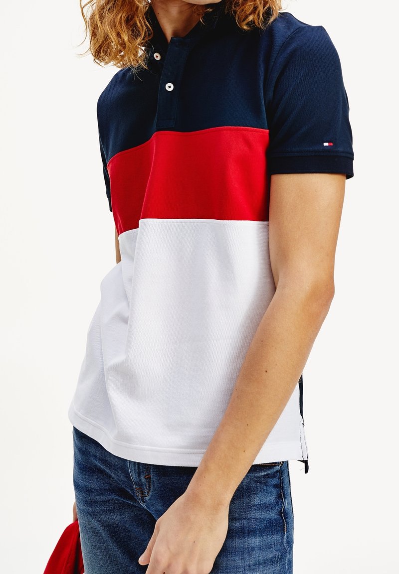 Personne portant un polo à manches courtes avec des blocs de couleurs horizontaux bleu marine, rouge et blanc, et un jean bleu, tenant un objet rouge dans la main gauche.