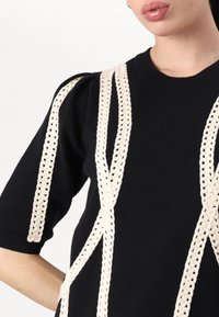 Zwarte gebreide top met crèmekleurige gevlochten details, korte mouwen en een ronde halslijn. Opmerkelijke textuur contrasteert met de gladde stof.