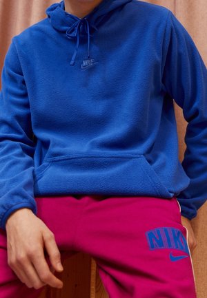 Osoba ubrana w niebieską bluzę z kapturem Nike z polaru oraz jaskrawo różowe spodnie dresowe Nike, siedząca z rękami spoczywającymi na udach.
