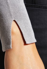 Světle šedý pletený svetr s rukávem s rozparkem blízko zápěstí, který ukazuje hladkou texturu látky a těsný střih.