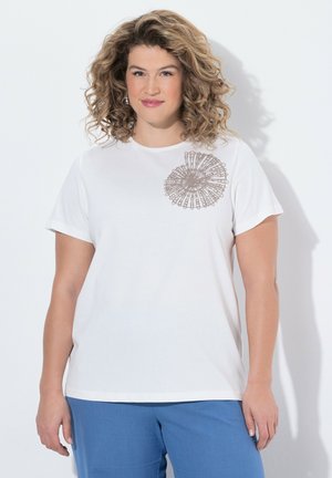 Frau mit lockigem Haar, die ein weißes T-Shirt mit einem kreisförmigen beigen Design auf der linken Brust und blaue Hosen trägt, steht vor einem weißen Hintergrund.