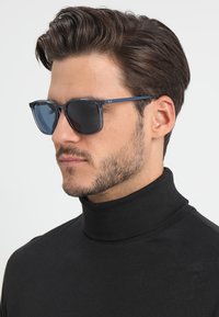 Ochelari de soare cu o ramă albastră închisă și lentile închise la culoare. Prezintă o formă rectangulară elegantă și o marcă minimală pe lateral.