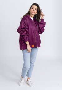 Oversized paarse bomberjack met geribbelde manchetten, gouden rits en oranje label. Gekocht met lichtblauwe jeans en witte sneakers.