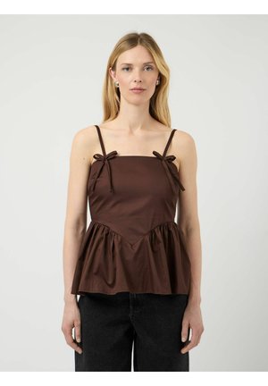 Donna bionda con un top marrone senza maniche con spalline sottili legate a fiocco e orlo arricciato, abbinato a pantaloni neri, in piedi su sfondo bianco.