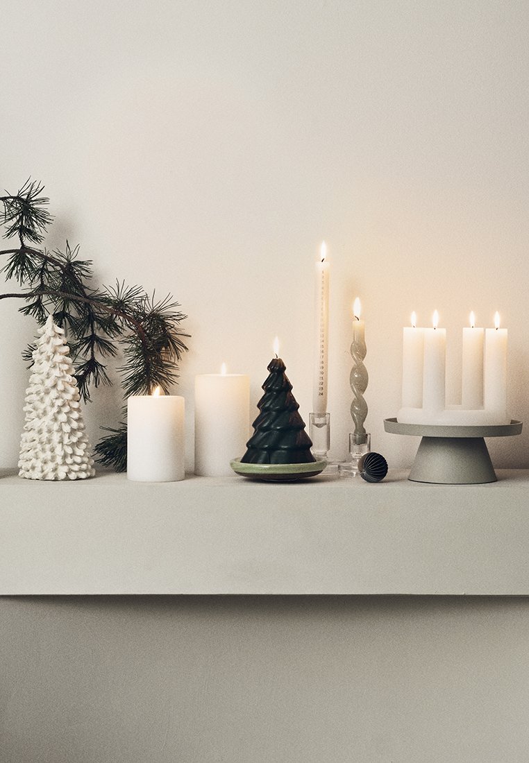 Kerzen in verschiedenen Formen und Höhen auf einem neutralen Regal. Zu den Merkmalen gehören ein strukturierter weißer Baum, eine grüne Baumkerze und glatte weiße Säulen.