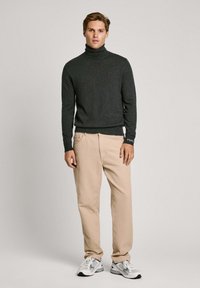 Maglione grigio scuro a collo alto con polsini a coste, abbinato a pantaloni dritti beige chiaro e sneaker grigie con accenti bianchi.