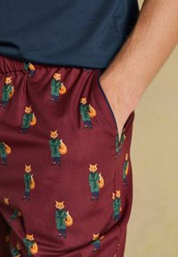 Tuvumā redzami bordo krāsas bikses ar atkārtojošos rakstu, kurā ir lapsas, kas ģērbtas zaļos mēteļos un zilās biksēs, viena roka izlīkusī kabatā.