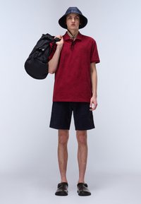 Röd polo med bladmönster, marinblå shorts, svart bucket-hatt och en svart duffelväska. Hållbara material och praktisk design.