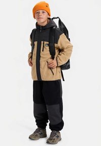 Ragazzo che indossa un berretto arancione, una giacca in fleece beige e nera, pantaloni neri, scarpe da trekking e porta uno zaino nero, in piedi con sicurezza.