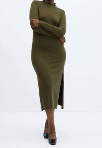 Femme portant une robe midi à manches longues vert olive avec une fente latérale et des escarpins Mary Jane noirs en cuir verni, posant devant un fond blanc.