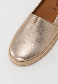 Metalické zlaté nazouvací boty s texturovaným povrchem, elastickým vrškem a pletenou podešví ve stylu juty. Vyznačují se čistými liniemi a minimalistickým designem.