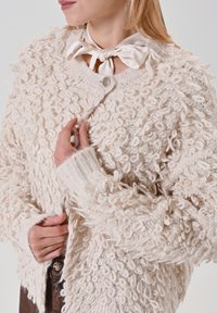 Cardigan beige crème texturé avec détails en tissu bouclé, doté d'une fermeture par bouton rond argenté et de poignets côtelés, porté sur un chemisier.