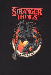 Černé tričko s podrobným grafikou tvora s ostrými drápy, obklopeného ohnivě oranžovým kruhem a textem "Stranger Things" nahoře.