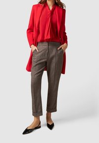 Manteau rouge, blouse rouge transparente avec nœud papillon, pantalon marron à carreaux, et ballerines pointues en cuir verni noir. Détails de tissu texturé visibles.