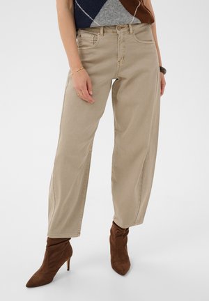 Person iført løse beige bukser og brune støvler med hæl, med en sweater med geometrisk mønster synlig foroven. Armene hænger afslappet ned langs siden.