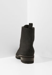 Botín negro hasta el tobillo con una superficie texturizada y brillante. Cuenta con una lengüeta, suela de goma con un ligero tacón y una silueta estilizada.