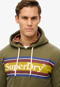 Sudadera verde oliva con bolsillo tipo canguro, con rayas multicolores en amarillo, burdeos y azul; logo de "SuperDry" exhibido de manera destacada.
