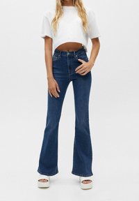 Džíny s vysokým pasem a rozšířenými nohavicemi v tmavém denimu s hladkou texturou, kombinované s krátkým bílým tričkem. Celkový vzhled doplňují sandály na platformě.