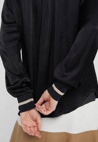 Schwarze Bluse aus strukturiertem Stoff, mit langen Puffärmeln und kontrastierenden goldgestreiften Manschetten. Der Saum ist hinten länger und liegt über einem weißen Oberteil.