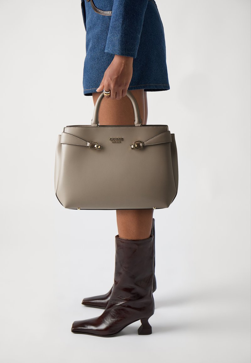Beige læder håndtaske med dobbelte top hanke, der har flade stropper og guldfarvede hardware detaljer, vist sammen med brune læder ankelstøvler.