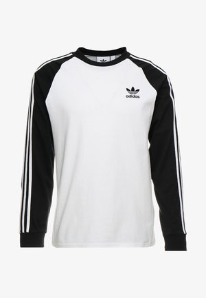 Pitkähihainen Adidaksen paita, jossa valkoinen runko, mustat hihat joissa on kolme valkoista raitaa, ja Adidas-logo vasemmassa rinnassa.