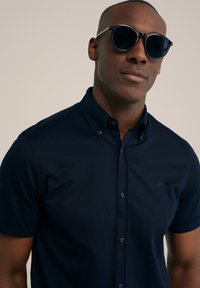 Polo de culoare bleumarin, cu mânecă scurtă, guler cu nasturi și un logo subtil. Modelul poartă ochelari de soare rotunzi, negri, evidențiind o textură netedă a țesăturii.