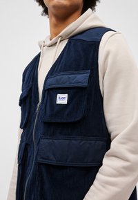Gilet in corduroy blu navy con zip frontale, diverse tasche grandi e una piccola etichetta con logo. Indossato sopra un felpa chiara.