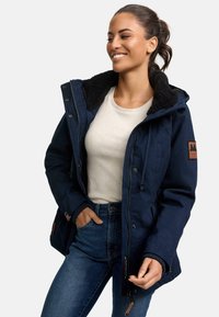 Navy blue jacket with a textured, black fleece-lined hood, brown leather patch detail, and elastic cuffs; worn over a light top and jeans.