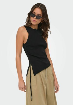 Femme portant un haut noir sans manches avec des liens sur les côtés, un pantalon beige à jambes larges, des lunettes de soleil noires cat-eye et des boucles d'oreilles hoop dorées, posant de profil.