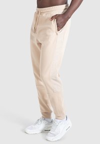 Beige joggingbroek van textuurstof, met een taps toelopend ontwerp, elastische tailleband, zijzakken en logo-detail, gecombineerd met witte sneakers.