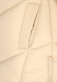 Nærbillede af beige quiltet jakke, der viser synede lommer og lynlåse detaljer på polstret stof.