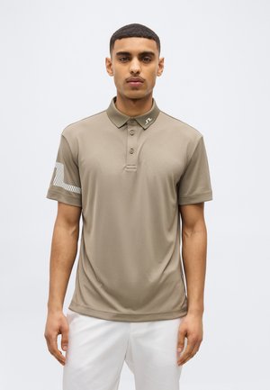 Mann trägt ein kurzärmeliges Poloshirt in Taupe mit einem Logo am Kragen und Ärmel, kombiniert mit weißen Hosen, steht vor einem einfarbigen Hintergrund.