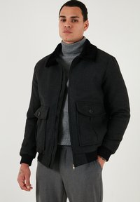 Črn bomberski jakna z mehkimi ovratnikom, zadrgo, dvema sprednjima žepoma, rebrastimi rokavi in trpežno teksturo tkanine. Nosi se preko sivega puloverja z visokim ovratnikom.