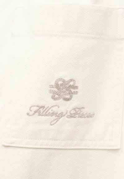 Λευκό ύφασμα με ραμμένη τσέπη, με μπεζ κεντημένο διαπλεγμένο λογότυπο πάνω από κείμενο με πλάγιους χαρακτήρες που γράφει "Filling Pieces".