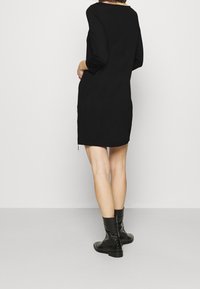 Femme portant une robe noire longueur genoux avec manches longues et des bottines noires, debout dos à la caméra.