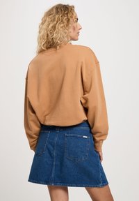 Luźny brązowy sweter z opadającymi ramionami oraz niebieska dżinsowa spódnica w kształcie litery A, wyposażona w dwie tylne kieszenie i widoczną metkę z logo.