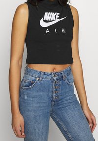Kvinna i ärmlös svart crop top med vit Nike Air-logotyp och högmidjade blå jeans med flera knappar, står mot en enfärgad bakgrund.