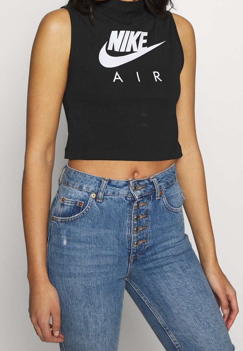 Kvinna i ärmlös svart crop top med vit Nike Air-logotyp och högmidjade blå jeans med flera knappar, står mot en enfärgad bakgrund.