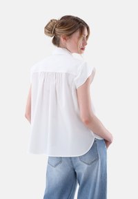 Weißes kurzärmliges Blusenshirt mit Kragen, ausgestattet mit einem gerafften Rückendesign. Kombiniert mit hellblauen weiten Jeans. Glatter, strukturierter Stoff.