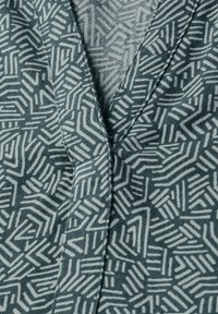 Wzorzysty materiał w odcieniach teal i jasnoszarym, z geometrycznymi wzorami zawierającymi linie i kształty oraz widocznymi przeszyciami na brzegach.