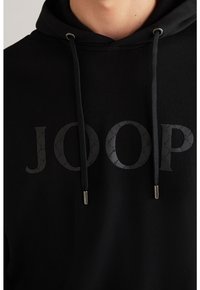 Sudadera negra con capucha que presenta un gran logo texturizado "JOOP" en gris oscuro. Incluye cordones con puntas metálicas y una textura de tela suave.