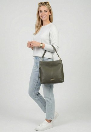 Mujer sonriente con suéter ligero y jeans, llevando un bolso de hombro de cuero verde oliva con etiqueta dorada, posando frente a un fondo liso.