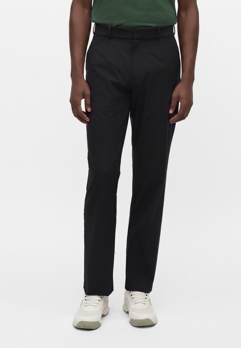 Personne portant un pantalon droit noir, une chemise verte rentrée et des baskets blanches, debout devant un fond blanc uni.