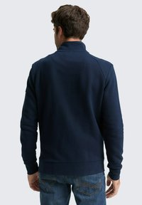 Marineblaues Sweatshirt mit hohem Kragen, strukturiertem Ripp und taillierten Bündchen. Der Rücken hat eine Nahtdetail und wird mit Jeans kombiniert.