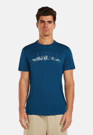 Man draagt een blauw T-shirt met een witte bergketen en gelabelde bergtoppen erop, gecombineerd met beige korte broek, staand tegen een effen achtergrond.