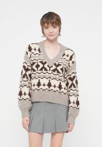 Pull en tricot crème et marron avec un col en V, présentant un motif géométrique. Design court et ourlet côtelé, associé à une jupe plissée grise.