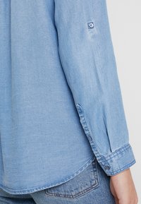 Light blue denim shirt with long sleeves, buttoned cuffs, and subtle texture. Hemline is slightly curved, creating a relaxed fit.