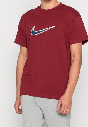 Persoon draagt een bordeauxrode T-shirt met korte mouwen met een donkerblauw en wit Nike Swoosh-logo, gecombineerd met lichtgrijze broek.
