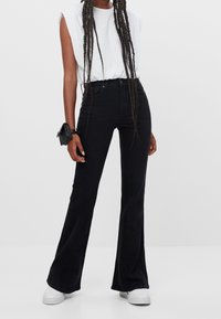 Schwarze schmal zulaufende Jeans aus Baumwoll-Denim mit hoher Taille und glatter Textur. Kombiniert mit einem weißen Kurzarmhemd und weißen Sneakers.